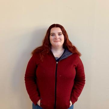 Beth, a white woman with red hair wearing a red zip up sweater, stands in front of a white wall and smirks at the camera
